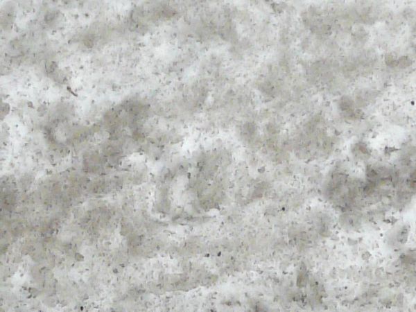 Road edge texture, with grey asphalt embedded with white rocks meeting edges of black-flecked snow, grey sidewalk and a white snow with grey patches.